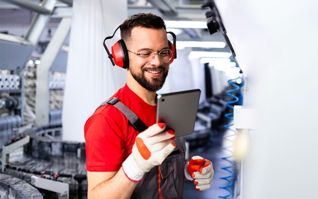 A factory worker wearing red ear protection and gloves uses a tablet device while standing in an industrial setting.