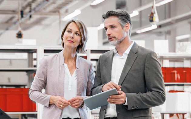 Two professionally dressed adults stand indoors; the man holds a tablet and gestures while the woman listens attentively. Shelves and industrial lighting appear in the background.