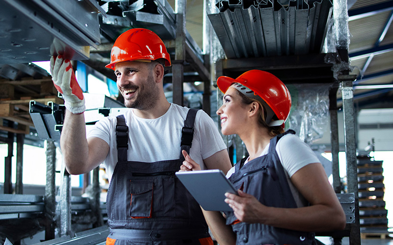 Two factory workers in overalls and orange helmets inspect metal structures while one holds a tablet in an industrial warehouse setting.