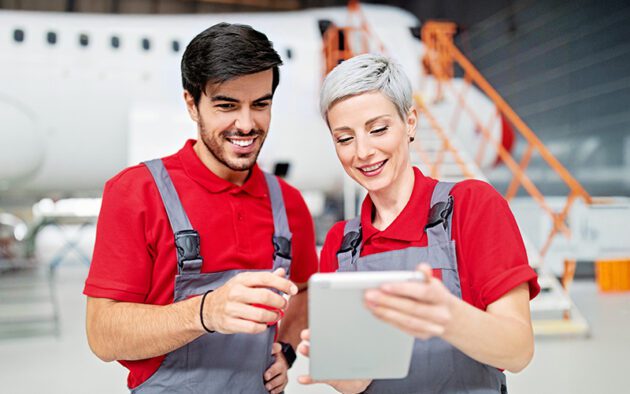 Two aircraft maintenance workers in red shirts and overalls look at a tablet together in a hangar with an airplane in the background.
