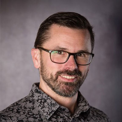 A man with short brown hair, a beard, and glasses, wearing a patterned shirt, smiles at the camera against a plain, dark background.