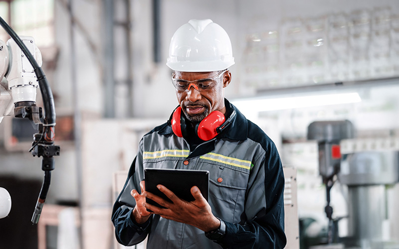 A factory worker wearing a hard hat, safety glasses, and ear protection uses a tablet in an industrial setting with machinery in the background.