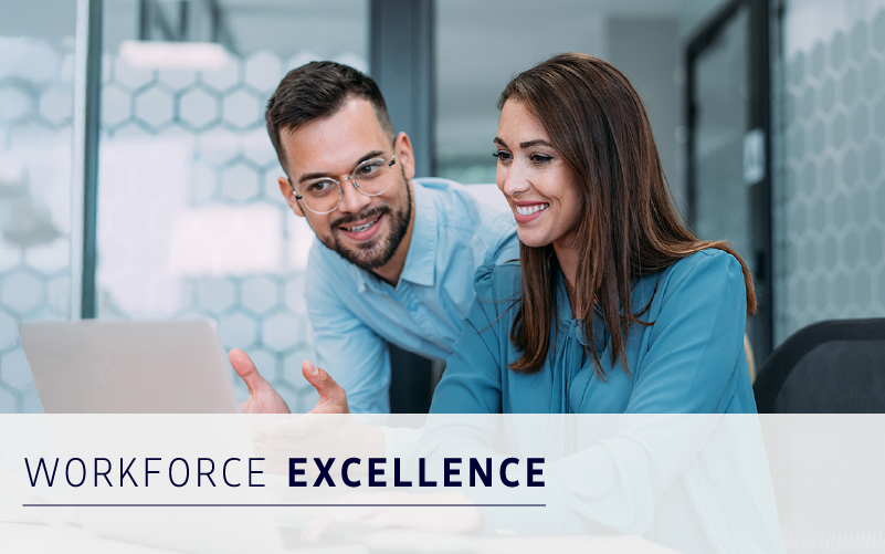 Two colleagues in business attire sit at a desk, smiling and looking at a laptop screen. Text overlay reads "Workforce Excellence.