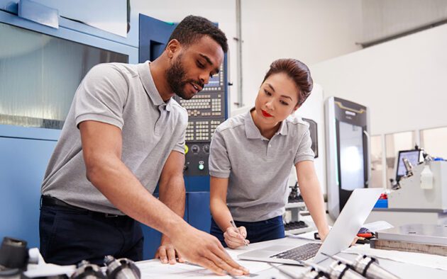 Two engineers in gray polo shirts review documents and data on a desk beside industrial equipment in a modern manufacturing facility.
