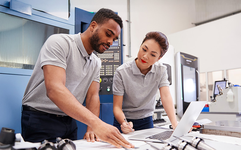 Two engineers in grey polo shirts review documents and work on a laptop in an industrial setting with machinery in the background.