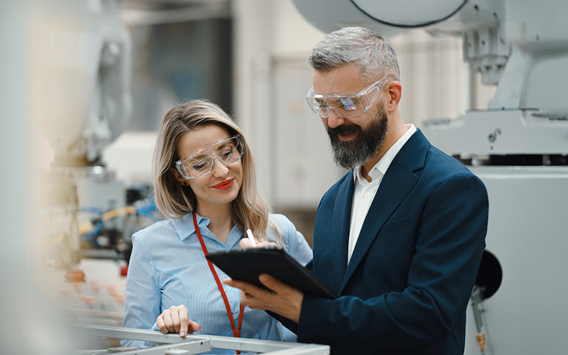 Two people wearing safety glasses stand in an industrial setting, looking at a clipboard and discussing information.