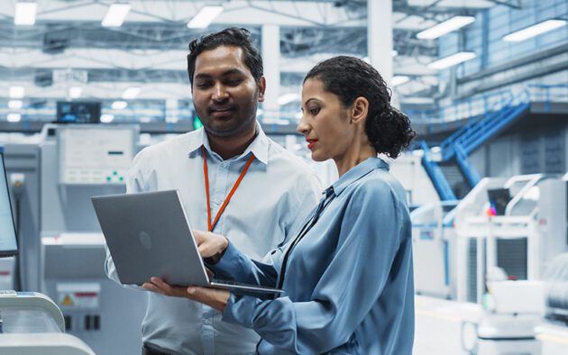 Two professionals in business attire stand in a modern industrial facility, reviewing information on a laptop computer.