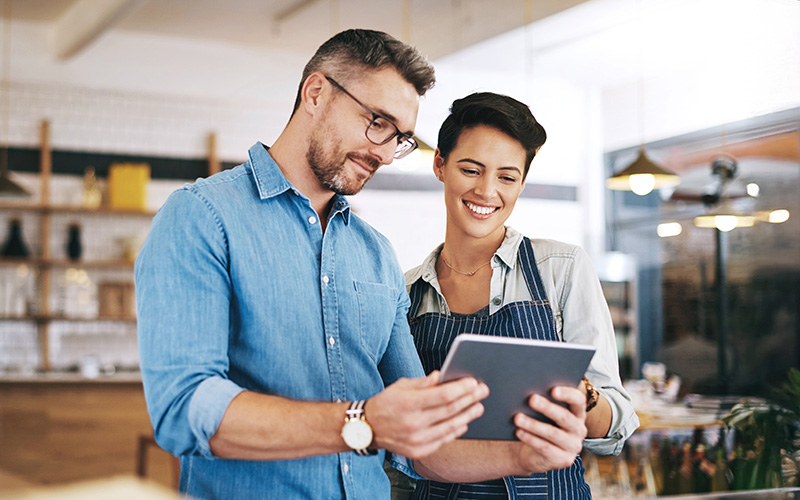 Two people stand indoors, looking at a tablet together; one wears glasses and a denim shirt, the other smiles in an apron as they discuss a workforce management solution.