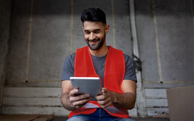 A man wearing a red safety vest sits indoors and uses a tablet, smiling as he looks at the screen.