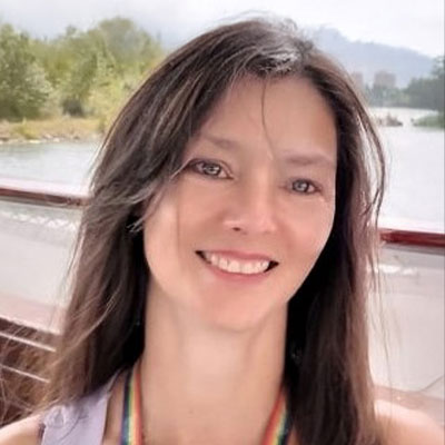 A woman with long brown hair smiles while sitting outdoors near water, with greenery and mountains visible in the background.