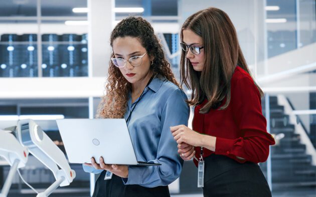 Two women in business attire look at a laptop together in a modern office setting with glass walls and visible stairs in the background.