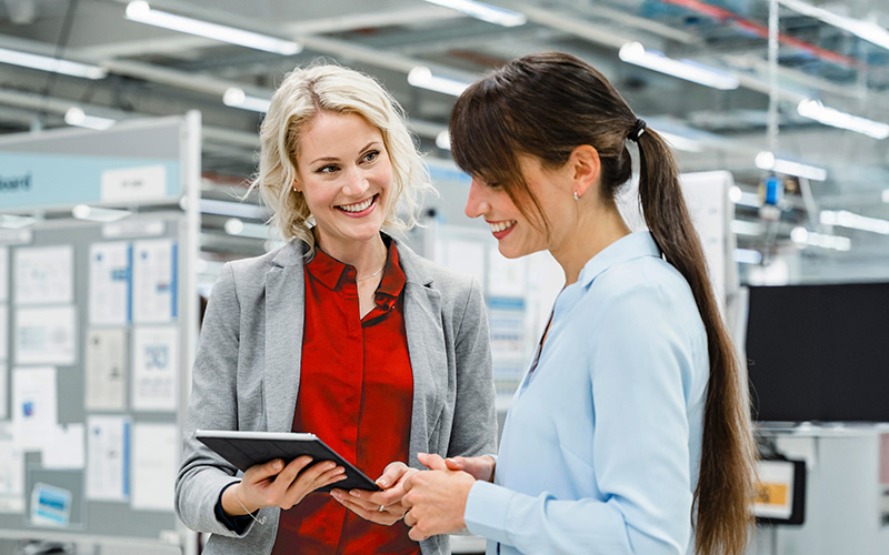Two women in business attire stand in an office or industrial setting, smiling and holding a digital tablet while having a discussion.