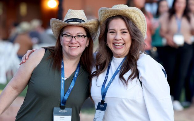 Two women wearing straw hats and conference name badges smile and pose together outdoors at an event.
