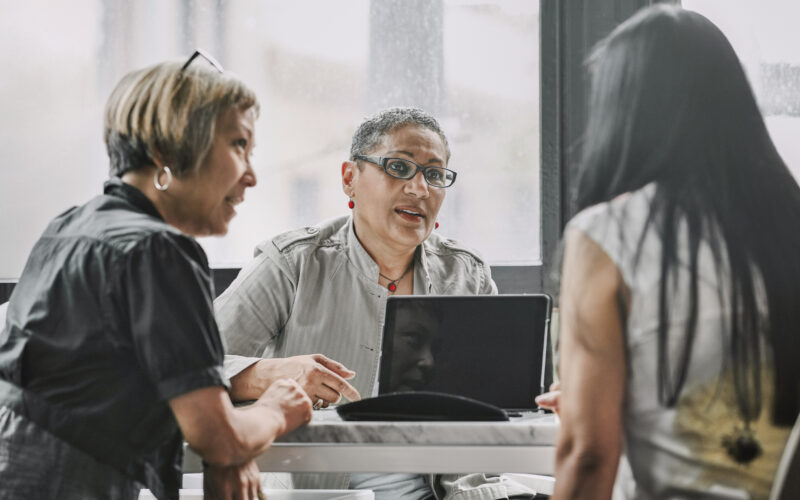 Three women sit at a table in discussion, with a laptop open in front of them, in a brightly lit room with large windows.