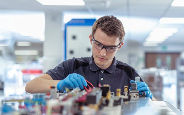 A person wearing safety glasses and gloves works with electronic components and wires on a laboratory bench.
