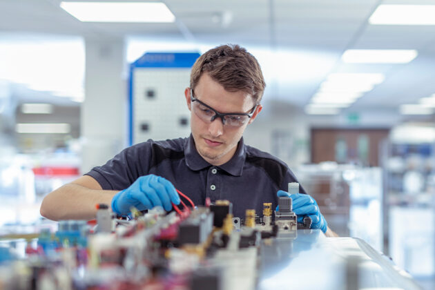 A person wearing safety glasses and gloves works with electrical components and wiring at a well-lit laboratory workstation.