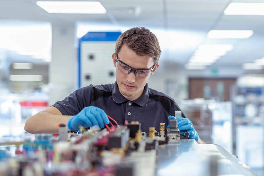A person wearing safety glasses and gloves works with electrical components and wiring at a well-lit laboratory workstation.