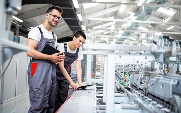 Two workers in gray overalls operate and inspect machinery on a factory production line in a well-lit industrial setting.