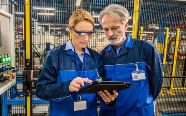 Two workers in blue uniforms and safety glasses review information on a tablet in an industrial facility.