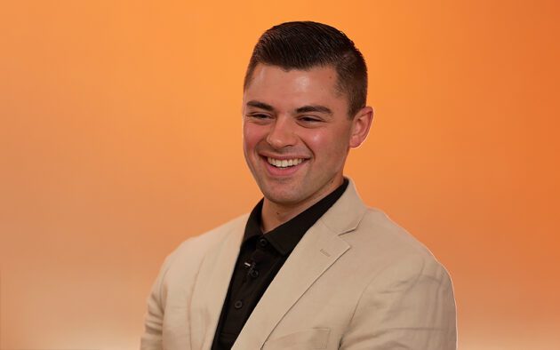 A man in a beige blazer and black shirt smiles in front of an orange background.