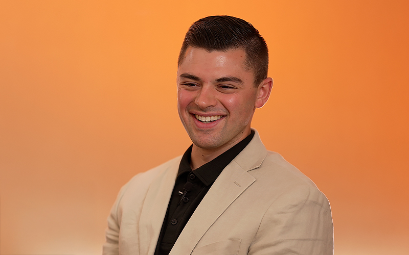 A man in a tan blazer and black shirt smiles against an orange gradient background.
