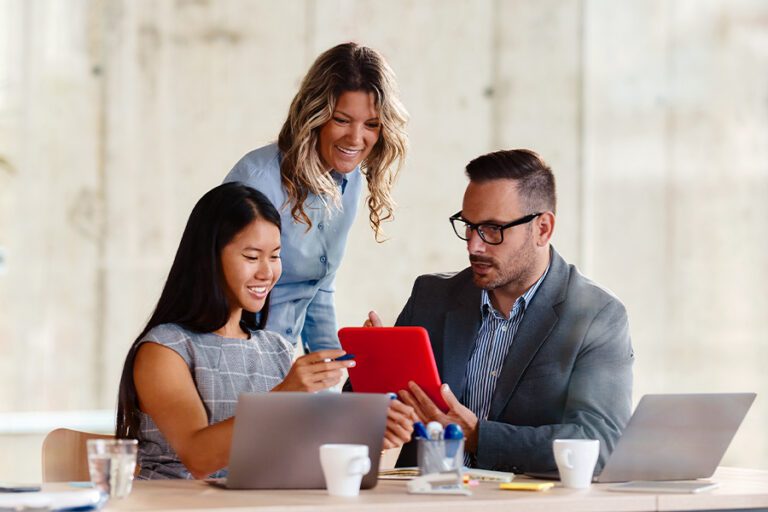 Three professionals collaborate at a table with laptops and coffee cups, as one person holds a red tablet and another points at the screen.