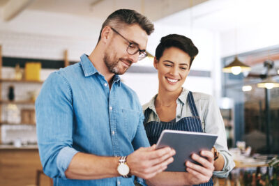 Two people standing indoors, looking at a tablet together; one person wears glasses and a denim shirt, the other wears an apron and smiles.