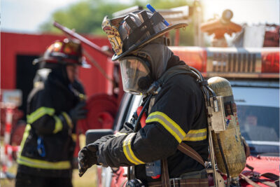A firefighter in full protective gear stands near a fire truck, adjusting gloves. Another firefighter is visible in the background.