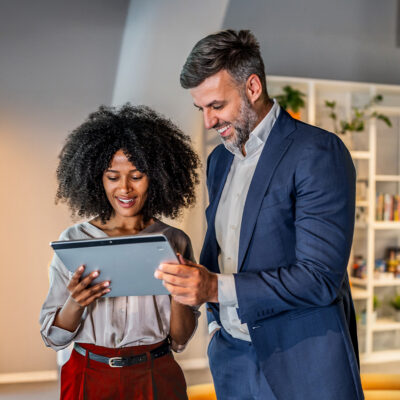 Two colleagues stand indoors, looking at a tablet together. The woman holds the device while the man in a suit gestures to the screen. Shelves with plants and books are in the background.