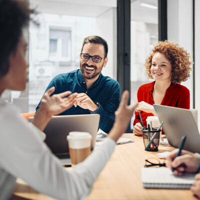 Three people sit at a table with laptops, coffee cups, and notebooks, having a discussion in a modern office setting.
