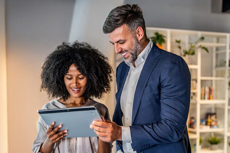 Two professionally dressed people review information together on a digital tablet in a modern office setting.