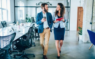 Two business professionals walk and talk in a modern office, one holding a coffee cup and the other holding a tablet, with empty desks and chairs in the background.