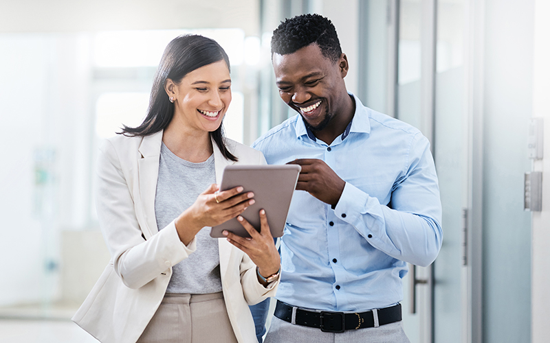 Two colleagues standing in a hallway, smiling and looking at a tablet together, both dressed in business attire.