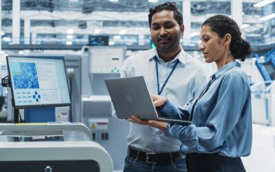 Two professionals in a factory setting review information on a laptop, standing beside industrial equipment and a monitor displaying technical data.