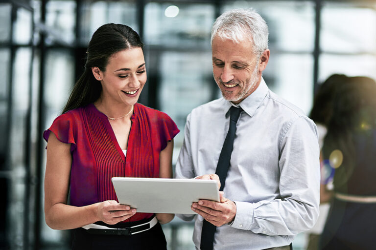 A woman and a man stand indoors, smiling and looking at a tablet together. The man wears a tie and shirt; the woman wears a red blouse.