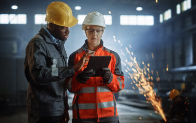 Two industrial workers wearing safety gear review information on a tablet in a factory, with sparks visible in the background.
