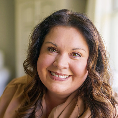 A woman with long brown wavy hair and a beige top smiles at the camera in a softly lit indoor setting, reflecting confidence often seen in successful compliance strategies.