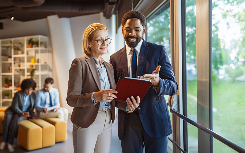 Two business professionals stand by a window, looking at a tablet and discussing something. In the background, two other people sit on a couch and talk.