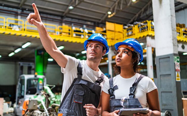 Two factory workers in blue helmets and overalls look in the same direction; one points while the other holds a digital tablet, highlighting effective workforce management inside a large industrial warehouse.