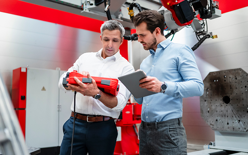 Two men in business attire examine a red industrial device while standing in a modern manufacturing facility; one holds a tablet.