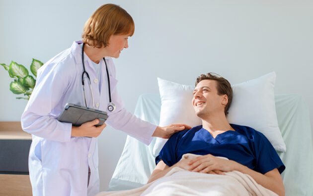 A doctor with a stethoscope talks to a smiling patient lying in a hospital bed, holding a tablet and placing a hand on the patient's shoulder.