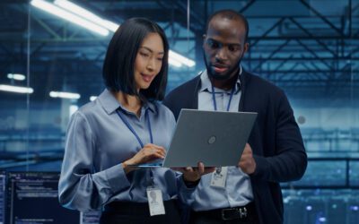 Two professionals wearing badges discuss Workforce Management while looking at a laptop together in a modern office with large windows and computer monitors in the background.