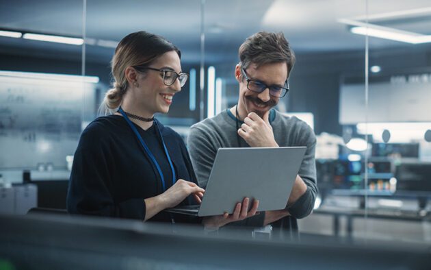 Two people in an office setting stand together looking at a laptop screen, discussing something and smiling. The environment appears modern and tech-focused.