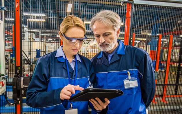 Two factory workers in blue uniforms examine information on a tablet in an industrial setting, with machinery and safety barriers in the background.
