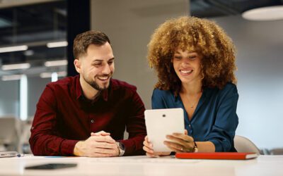 Two people sit at a table in an office, smiling and looking at a tablet device together.