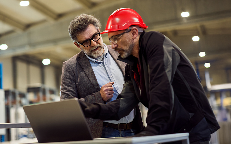 Two men, one in a suit and glasses and the other in a red hard hat and workwear, discuss something while looking at a laptop in an industrial setting.