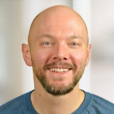 A smiling bald man with a trimmed beard and mustache wears a blue shirt and is standing in front of a blurred light background.