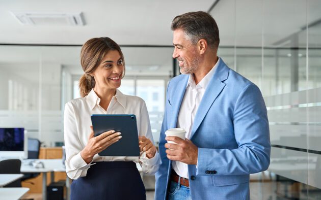 Two professionals in business attire stand in an office, smiling and conversing; one holds a tablet and the other holds a coffee cup.