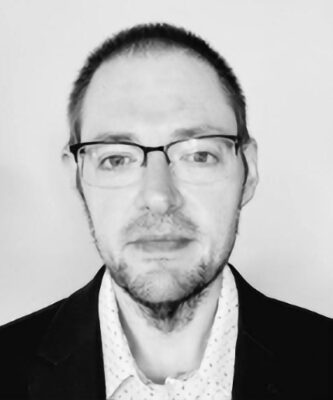 A man with short hair, glasses, and a beard wearing a dark blazer and a light-colored shirt, facing the camera against a plain background.