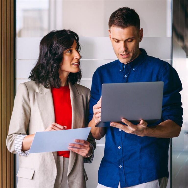 Two colleagues stand indoors discussing something; the woman holds documents while the man gestures at a laptop screen.